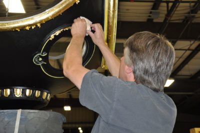 "An artisan is shown in the process of applying gold leaf to the ornate trim of a street clock in this photo. With each piece of gold leaf carefully applied, the clock's trim begins to shimmer and shine, highlighting its intricate design and drawing attention to its beauty. The artisan's skillful application of the gold leaf ensures that the clock's trim is transformed into a work of art that catches the eye and adds a touch of elegance to the streetscape."